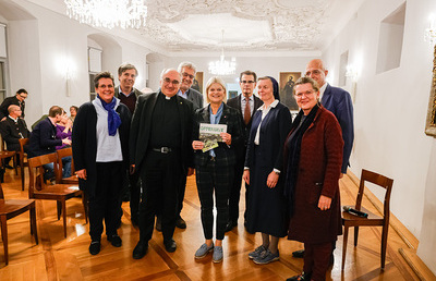 Verteidigungsministerin Klaudia Tanner mit der letzten Ausgabe der 'Offensive' der Katholischen Aktion zum Thema 'Frieden', und Bischof Wilhelm Krautwaschl, umringt von Vertreter:innen der einzelnen Kurien des KLRÖ